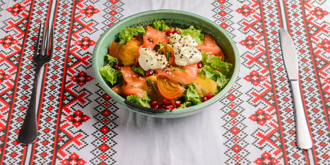 Delicious healthy salmon salad with cheese and pomegranate seeds served in a bowl over traditional Ukrainian tablecloth.
