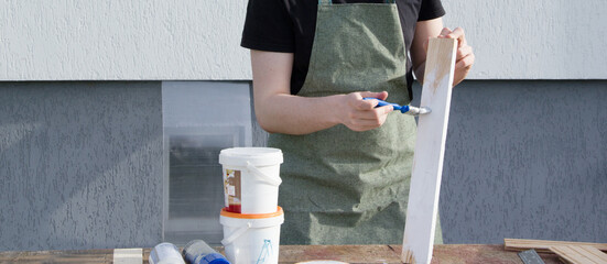 A young male designer paints a wooden Board white with a brush. The young man is working near his house. 