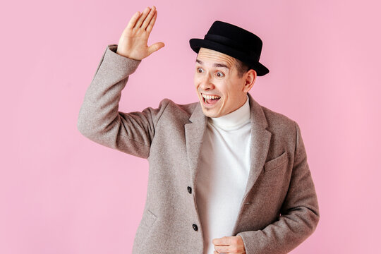 Saluting emotional man dressed in a grey beige coat and a classical porkpie hat waving his hand to someone. Over dark pink background.