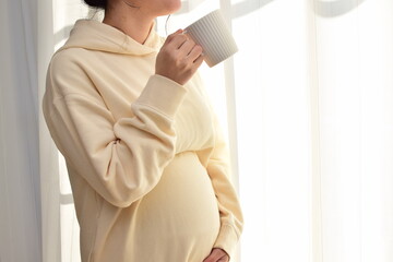 A pregnant woman in her twenties is drinking a drink in a mug. 