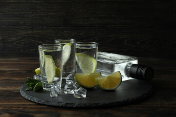 Tray with bottle and shots of vodka, lime and ice on wooden background