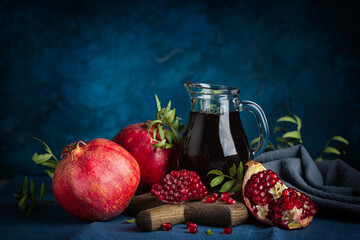 Pomegranate juice in a jug, whole pomegranates and seeds on dark blue background. Dark and moody.