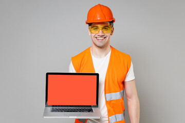 Young employee man in orange vest protective helmet hold laptop pc computer, blank screen workspace area isolated on grey background studio Instruments for renovation apartment Repair home concept