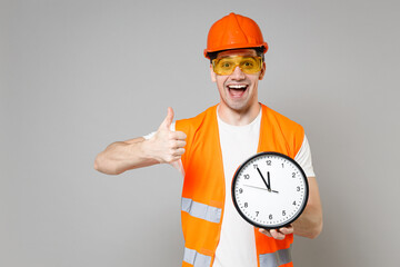 Young employee man in orange vest protective helmet hold in hands clock show thumb up gesture isolated on grey background studio portrait Instruments for renovation apartment Repair home concept