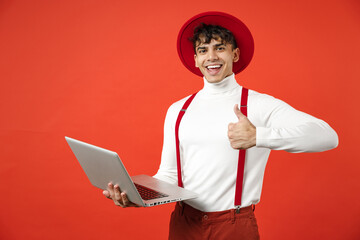 Young spanish latinos smiling happy confident fashionable freelancer man 20s wear hat, white shirt hold laptop pc computer show thumb up like gesture isolated on red color background studio portrait.