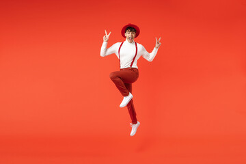 Fototapeta na wymiar Full length of young spanish latinos overjoyed smiling fun happy man in hat white shirt trousers with suspenders jumping high show v-sign victory gesture isolated on red background studio portrait.
