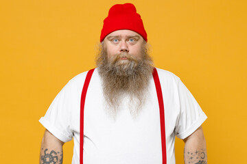 Fat pudge obese chubby overweight caucasian blue-eyed bearded tattooed man 30s has big belly wear white t-shirt red hat suspenders looking camera isolated on yellow color background studio portrait.