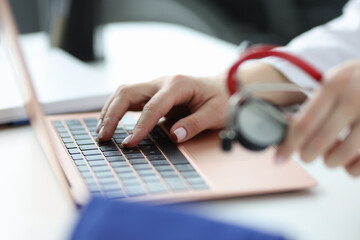 Doctor holding stethoscope and typing on laptop keyboard closeup