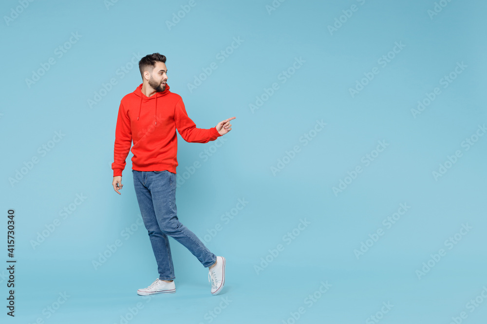 Wall mural Full length of young caucasian smiling bearded man 20s ?? casual red orange hoodie pointing on workspace area copy space mock up isolated on blue background studio portrait People lifestyle concept.