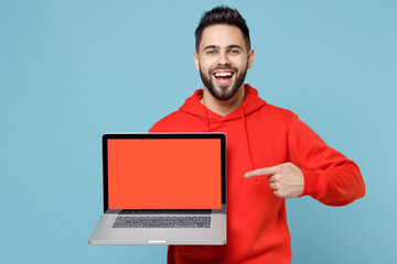 Young caucasian smiling web designer bearded man 20s in casual red orange hoodie point index finger on laptop pc computer with blank screen workspace area isolated on blue background studio portrait.