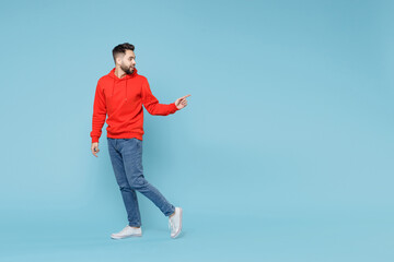 Full length of young caucasian smiling bearded man 20s ?? casual red orange hoodie pointing on workspace area copy space mock up isolated on blue background studio portrait People lifestyle concept.