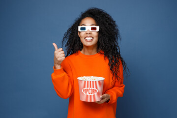 Funny young african american woman in casual bright orange sweatshirt 3d glasses watching movie film, hold bucket of popcorn showing thumb up like gesture isolated on blue background studio portrait.