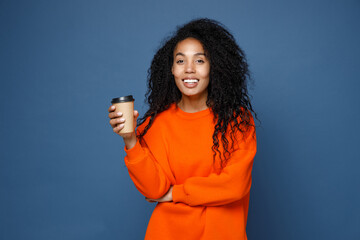 Smiling beautiful young african american woman wearing casual basic bright orange sweatshirt standing hold paper cup of coffee or tea looking camera isolated on blue color background studio portrait.