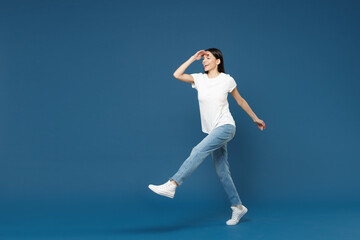 Obraz premium Full length young smiling latin woman 20s wearing white casual basic t-shirt holding hand at forehead looking far away distance raised up leg for step isolated on dark blue background studio portrait.