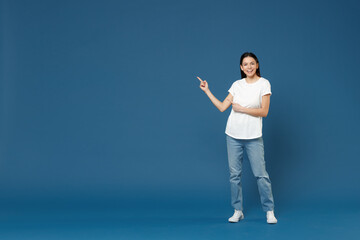 Full length young smiling latin woman 20s wearing white casual basic t-shirt pointing index finger aside back on copy space mock up area workspace isolated on dark blue background studio portrait.