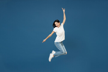 Full length side view of young happy sport smiling brunette latin woman 20s wear white casual basic t-shirt hold hand outstretched jumping high isolated on dark blue color background studio portrait.