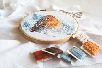cross stitch process in the hoop multicolored bird, white canvas, floss threads on bobbins, needle, needlework and cross stitch concept