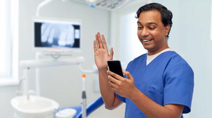 Naklejka na ściany i meble medicine, dentistry and technology concept - happy smiling indian doctor or male dentist in blue uniform having video call on smartphone over dental clinic background