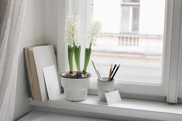 Easter spring still life. Blank greeting and businesss card mockups. Old books on window sill. White hyacinth in flower pot. Pencils in ceramic holder. Home office concept. Scandinavian interior