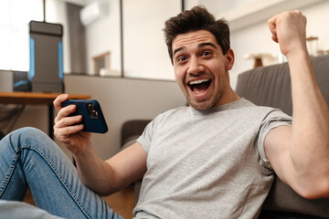 Delighted guy making winner gesture while playing video game on cellphone