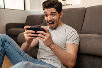 Delighted brunette guy playing video game on mobile phone at home