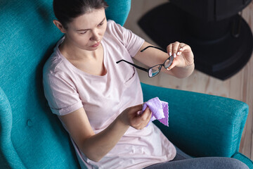 Young woman cleaning eyeglasses with a napkin while sitting in a chair