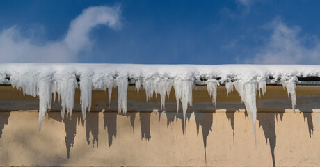 Snow and ice icicles on the roof of the building. Frozen water in white icicles. Contrasting shadows. Panoramic view. Blue sky with clouds.