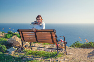 Young beautiful girl traveler with white jacket looking at camera, sit and smile on bench at Dingli Cliffs with Mediterranean sea blue water background, tourist woman on vacation in Malta island