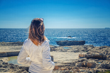 Young woman traveler with white jacket looking away distance at horizon of Mediterranean sea at coast of Dwejra Bay with stone rocks and cliffs of Gozo island, Malta in summer day with blue sky
