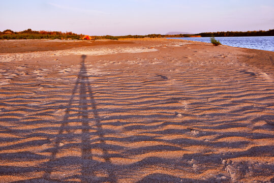Shadow of a tripod with a camera on the background of the harmonious lines of a sandy river bank at sunset