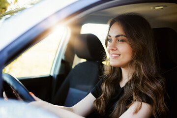 Beautiful young modern girl driving a car on sunny summer day. Female driver alone in the car, close-up, sun on the background.