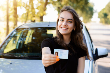 Pretty girl with cheerful smile standing near the car and showing driving license to the camera. Woman expresses her happiness after passing the driving exam. - obrazy, fototapety, plakaty