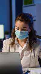 Employee with protection face mask working late at night in new normal business office to respect deadline of project, taking notes, analysing documents sitting at desk overtime during global pandemic