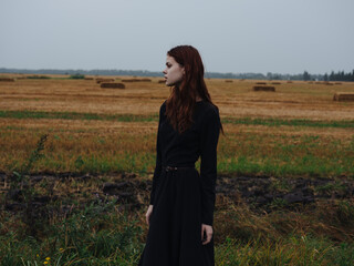 Obraz premium Beautiful woman in a black dress with red hair and a dry field in the background