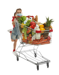 Little girl in medical mask with shopping cart full of groceries on white background