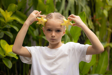 Charming little girl with two slices of fresh starfruit. Caucasian girl holding carambola in her hands. Selected focus. Green tropical leaves background. Organic fruit concept.