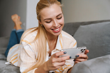 Happy blonde woman smiling and using cellphone while lying on couch