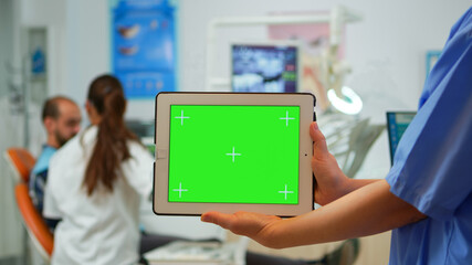 Fototapeta na wymiar Close up dentist nurse holding tablet with greenscreen display standing in stomatologic clinic, while doctor is working with patient in background. Using monitor with chroma key izolated pc key mockup