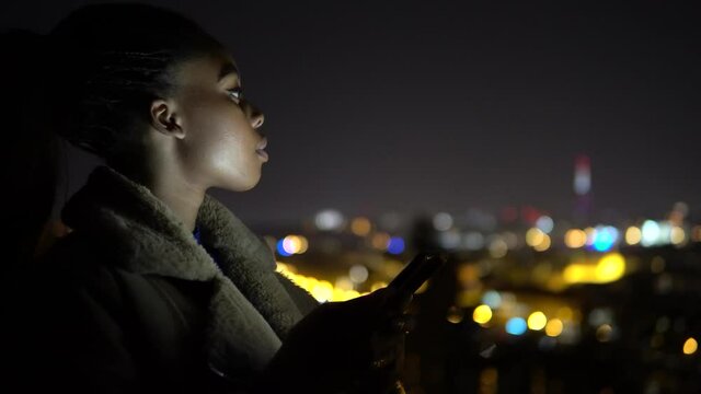 A young black woman works on a smartphone and looks around in an urban area at night - side closeup - city lights in the blurry background