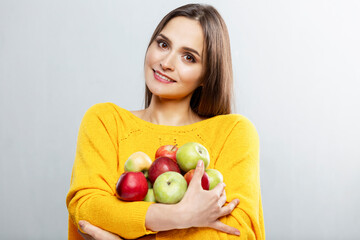 Young smiling woman holding many red and green apples in her hands. Beautiful brunette in a yellow sweater. Healthy food, vitamins and vegetarianism. Grey background.