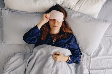 A young woman in a silk sleepwear sleeping in a bed with eyes covered with mask.