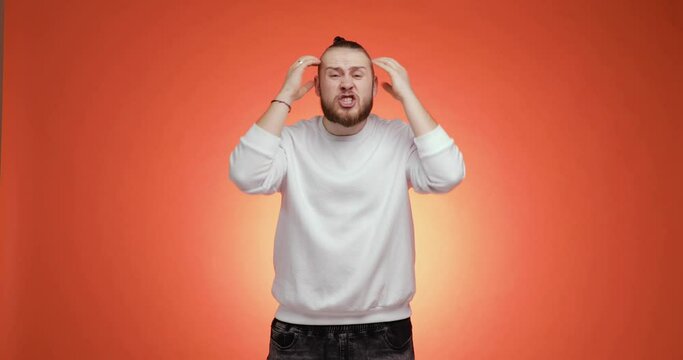 Surprised confused bearded young man with hair tail saying what the fuck on red background. He grabs his head and swears. People sincere emotions lifestyle concept. Wearing white sweater and jeans.
