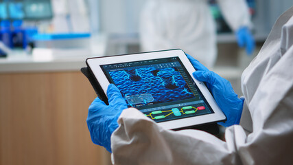 Back view of doctor in protection suit analyzing virus evolution looking on digital tablet. Team of...