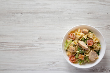 Homemade Chicken Caesar Pasta Salad in a white bowl on a white wooden surface, top view. Flat lay, overhead, from above. Copy space.