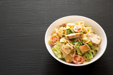 Homemade Chicken Caesar Pasta Salad in a white bowl on a black surface, side view. Copy space.