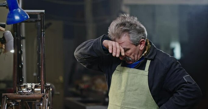 Tired elderly mechanic wiping sweat. Exhausted senior man with mustache wiping sweat from forehead and smiling for camera then starting to repair metal mechanism in workshop