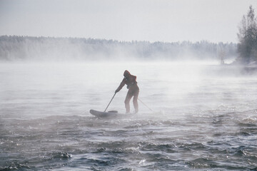 Surfer in a red wetsuit on a board with a paddle floats against the waves. Winter sup surfing on the Vuoksa river. Losev rapids. © AndreyZayats