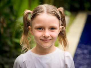 Close up portrait of pretty little girl. Blue eyes and blond hair. Caucasian girl with two ponytails. Green tropical leaves and swimming pool background.