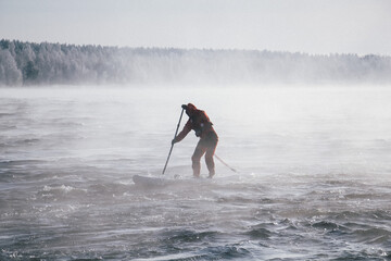 Surfer in a red wetsuit on a board with a paddle floats against the waves. Winter sup surfing on the Vuoksa river. Losev rapids. © AndreyZayats