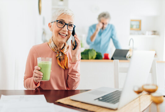 Senior Woman Talking On The Phone, Drinking Smoothie And Using Laptop While Man Cooking In Kitchen